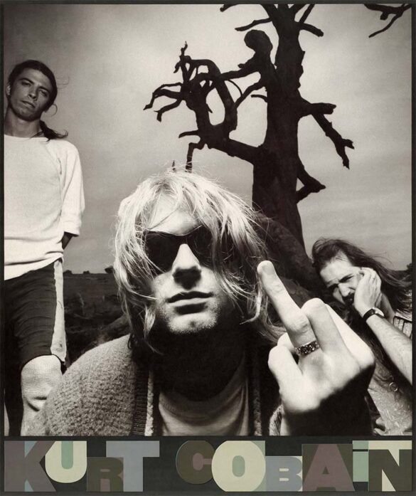 Black-and-white 1994 photo of Nirvana with Kurt Cobain in sunglasses and a cardigan in the foreground, giving the ring finger, while Dave Grohl and Krist Novoselic stand behind him near a bare tree.