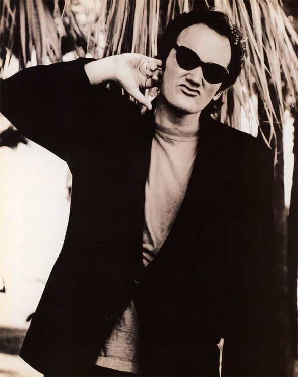 Black-and-white portrait of Quentin Tarantino wearing dark sunglasses and a jacket, posing outdoors with one hand raised near his face.