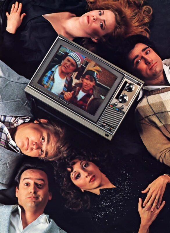 1982 color portrait of John Candy, Eugene Levy, Catherine O’Hara, Rick Moranis, Dave Thomas, Joe Flaherty, and Andrea Martin posed around a portable television.