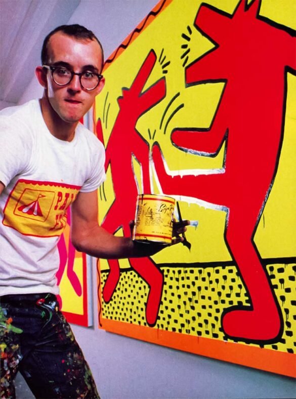 Keith Haring standing beside a large red and yellow painting, wearing glasses and paint-splattered pants while holding a paint can.