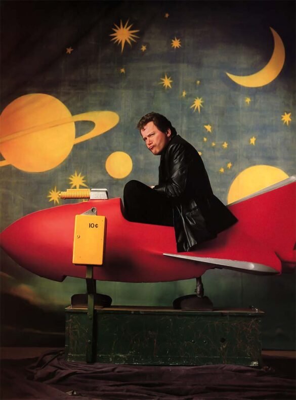 William Shatner seated on a red coin-operated rocket ride in front of a painted outer-space backdrop with planets, stars, and a crescent moon.
